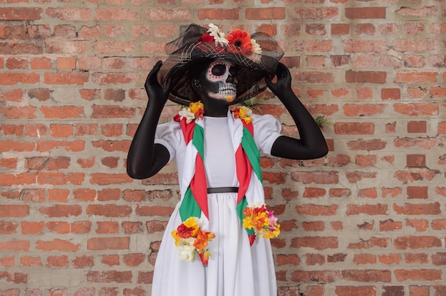 Front view of a woman in a costume of Calavera Catrina holding with both hands her black hat