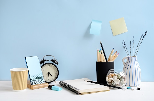 Front view of workspace with notepad, phone, pencils, coffee, clock on white table over blue wall. Mockup with copy space