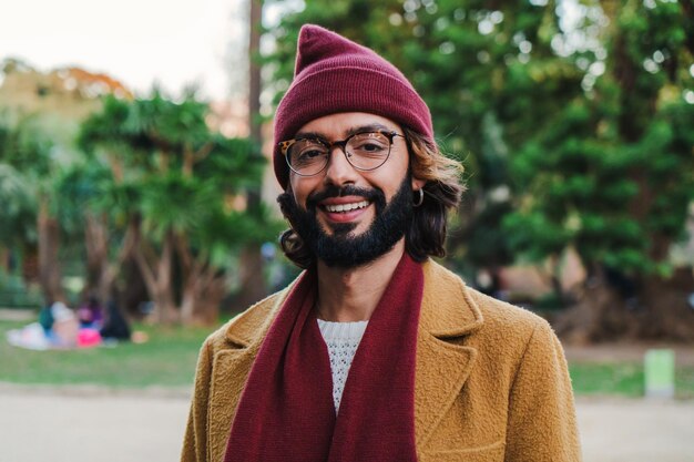 Front view of young adult man with goggles and beard smiling and looking at camera wearing a beanie hat and scarf in a park outdoors with happy and sucessful attitude Portrait concept