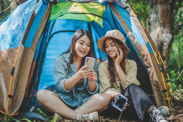 전면 보기 텐트 앞에 앉아 있는 젊은 아시아 예쁜 여자와 그녀의 여자 친구는 휴대 전화를 사용하여 함께 숲에서 캠핑하는 동안 행복과 함께 사진을 찍는다
