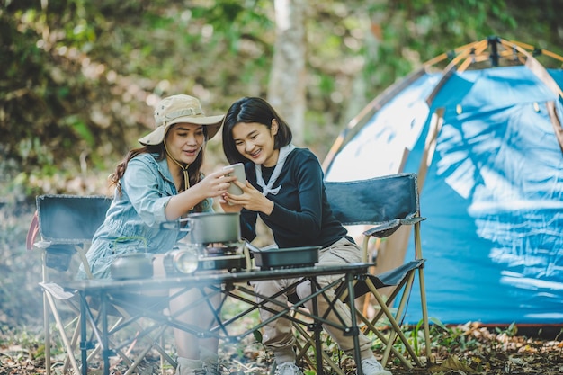 Front view Young Asian pretty woman and her girlfriend sitting at front of tent use mobile phone take photo during camping in forest with happiness together