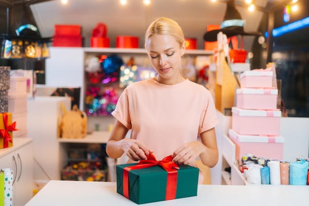 Front view of young attractive sales woman wrapping christmas gift box tying red ribbon and