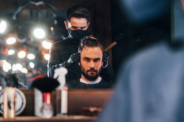 Front view of young bearded man that sitting and getting haircut in barber shop by guy in black protective mask