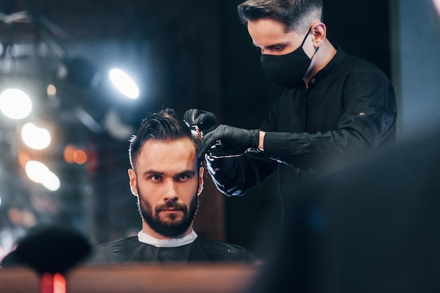 Front view of young bearded man that sitting and getting haircut in barber shop by guy in black protective mask