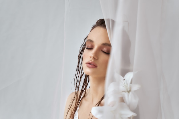 Front view of young brunette woman with perfect makeup, clean skin and closed eyes holding lily flower. Portrait of girl with wet hair posing on white background between tulle. Concept of beauty.