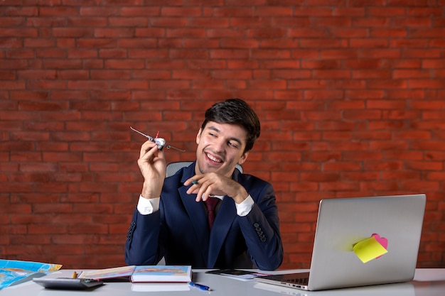 front view of young businessman sitting behind his working place in suit holding toy airplane work plan contractor business job builder corporate project