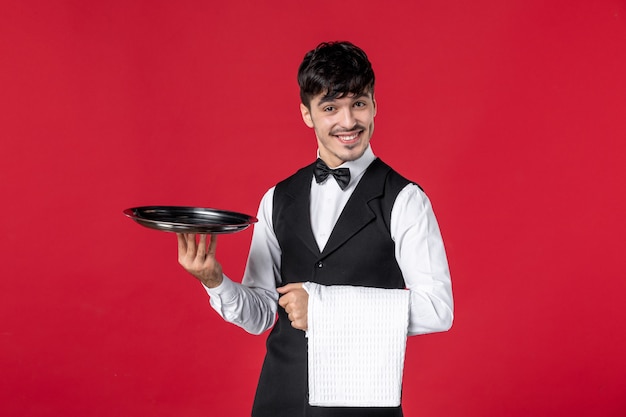 Front view of young confident male waiter in a uniform with butterfly on neck holding tray and towel on red wall