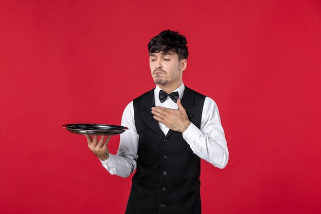 Front view of young dissatisfied man waiter in a uniform tying butterfly on neck holding tray on isolated red wall