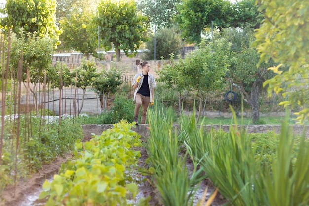 Вид спереди на молодого фермера с мотыгой и свежим фруктовым садом впереди с множеством разных овощей