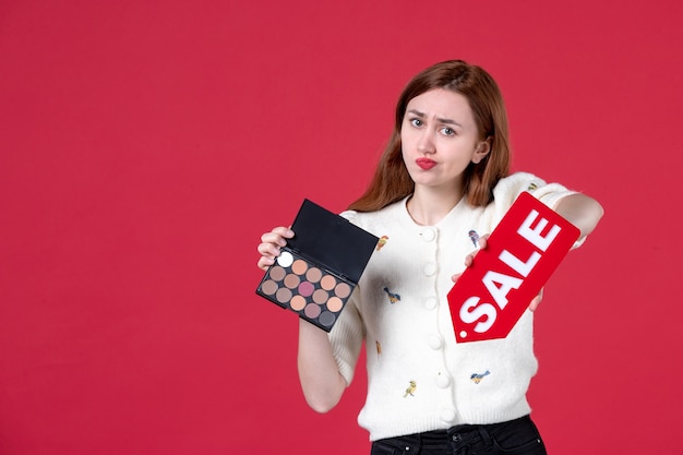 front view young female holding powder-box and red sale nameplate on red background sensual shopping woman color march make-up beauty fashion lips