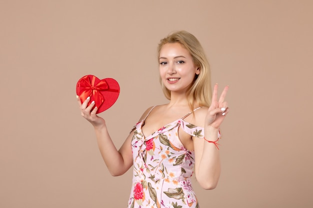 Front view of young female posing with red heart shaped present on brown wall