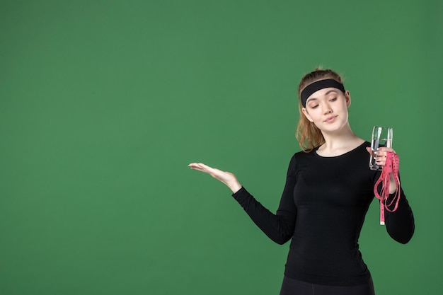 Vista frontale giovane donna con bicchiere d'acqua e misura della vita su sfondo verde colore corpo nero allenamento donna atleta sport