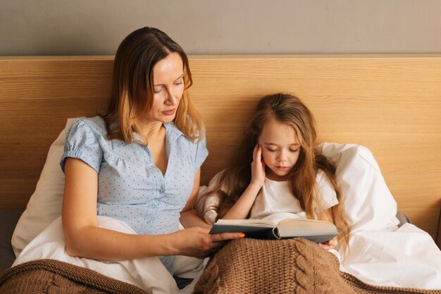 Front view of young happy mother and adorable cheerful daughter reading children book together lying in bed before going to sleep. Concept of family leisure activity at home.