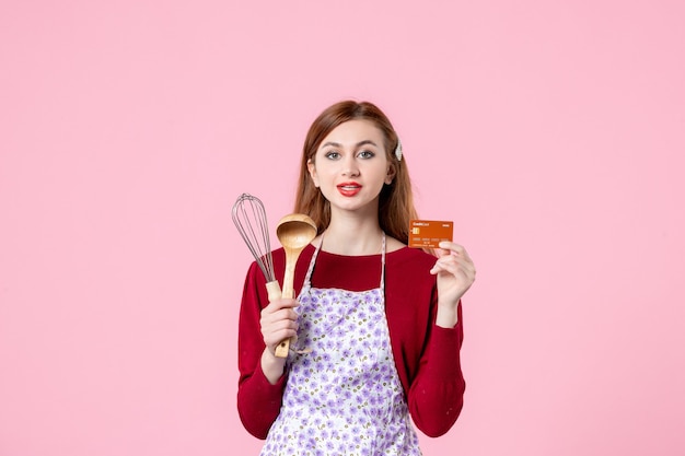 Foto vista frontale giovane casalinga che tiene frusta e carta bancaria su sfondo rosa torta donna colore torta cucina cucina denaro cucina cibo