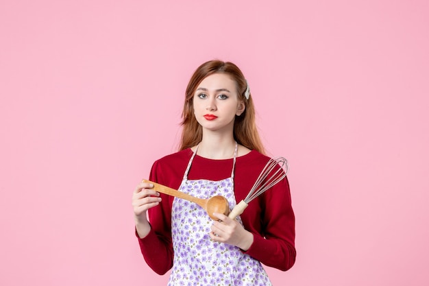 front view young housewife holding whisk and wooden spoon on pink background cake cuisine profession horizontal uniform dough woman cooking pies