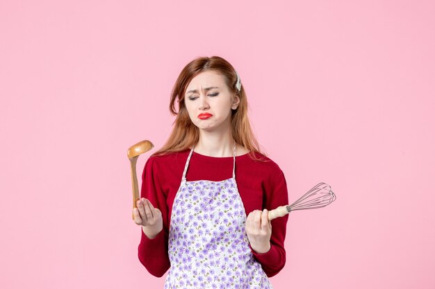 ピンクの背景の職業水平均一ケーキ生地の女性料理パイに泡立て器と木のスプーンを保持している正面図若い主婦