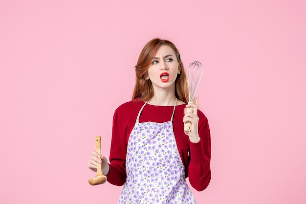 front view young housewife holding whisk and wooden spoon on pink background woman cooking horizontal pie cake uniform profession