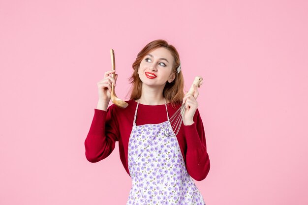 正面図ピンクの背景に泡立て器と木のスプーンを保持している若い主婦女性料理水平パイケーキ制服を調理
