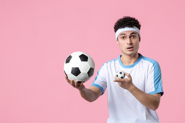 Vista frontale giovane atleta maschio in abbigliamento sportivo con pallone da calcio sulla parete rosa