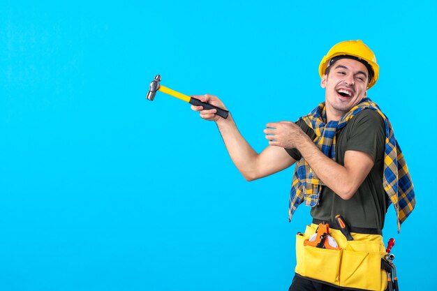 front view young male builder holding hammer laughing on the blue background house job flat building worker architecture constructor