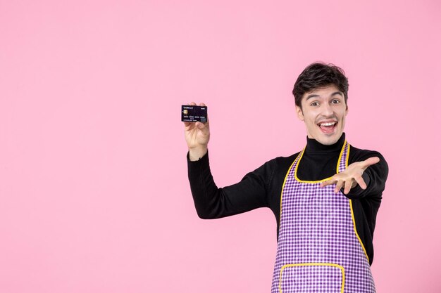 Vista frontale giovane maschio in mantello che tiene carta bancaria su sfondo rosa professione orizzontale lavoratore uniforme cucina colore lavoro