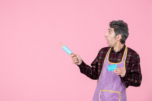 front view young male in cape holding little rolling pin and bank card on pink background husband worker uniform money horizontal profession kitchen cooking