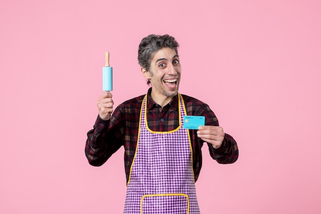 front view young male in cape holding little rolling pin and bank card on pink background profession husband horizontal kitchen money cooking job worker