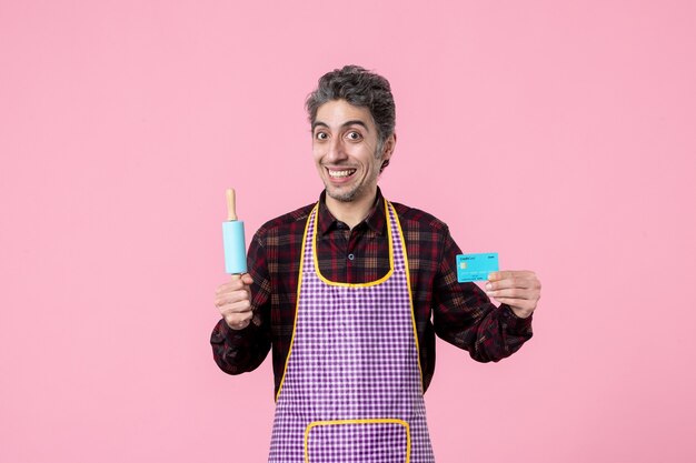 front view young male in cape holding little rolling pin and bank card on pink background profession husband horizontal kitchen uniform money cooking worker