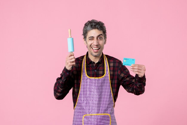 front view young male in cape holding little rolling pin and bank card on pink background profession husband worker horizontal kitchen money cooking job