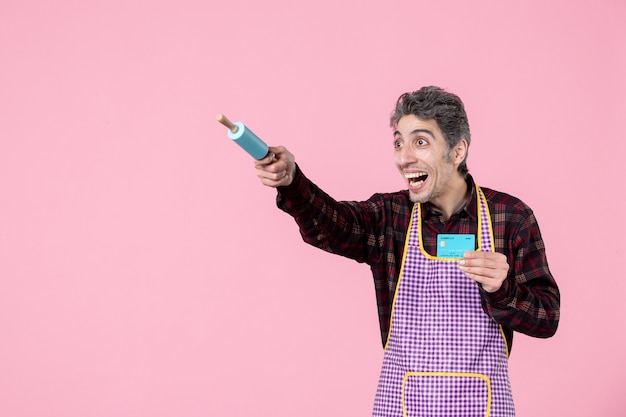 front view young male in cape holding little rolling pin and bank card pointing on pink background job worker kitchen cooking profession horizontal uniform money husband