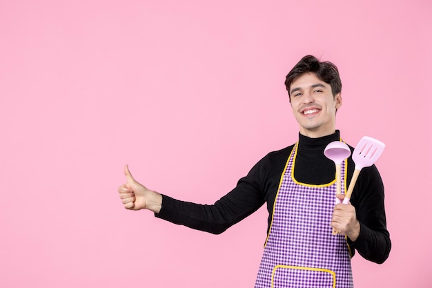 Вид спереди молодой мужчина в плаще держит розовые ложки на розовом фоне тесто профессия кухня униформа цвет горизонтальный главный кулинар