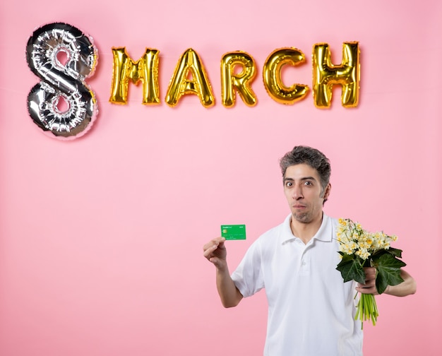 front view young male holding flowers for woman and bank card with march decorated pink background sensual womens day equality glamour feminine holiday party