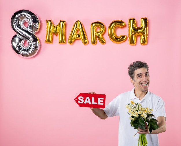 front view young male holding flowers for woman and sale nameplate with march decorated light-pink background equality glamour party shopping feminine