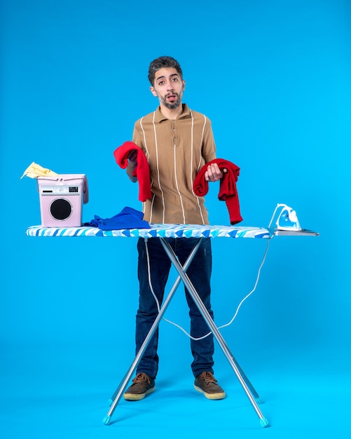 front view young male holding red clothes behind ironing board on blue background color housework laundry man emotion clean wash machine