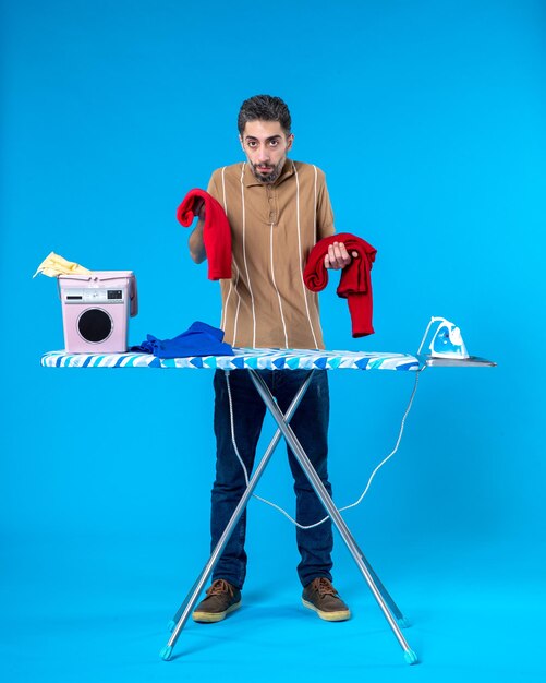 front view young male holding red clothes behind ironing board on the blue background color housework laundry man emotion clean wash machine