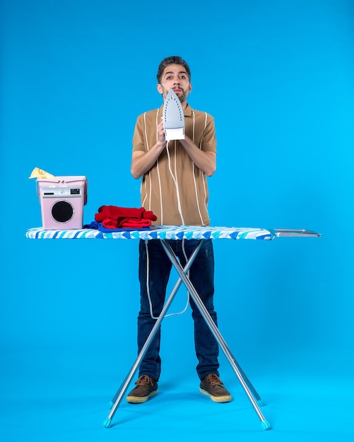 front view young male ironing behind board on the blue background color housework clean laundry man machine wash emotion