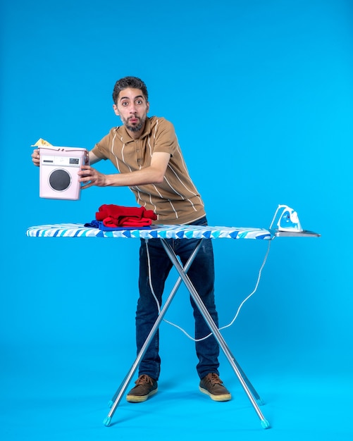 front view young male behind ironing board on a blue background color housework laundry man emotion clean wash machine