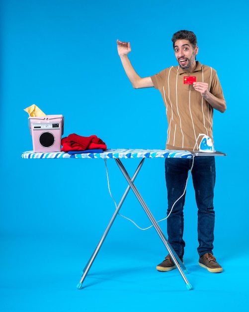 front view young male behind ironing board holding bank card on blue background clean laundry housework washing machine money clothes color