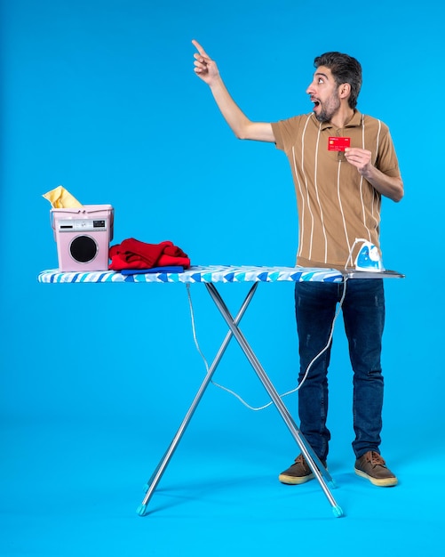 front view young male behind ironing board holding bank card on blue background laundry housework washing machine iron money clean color