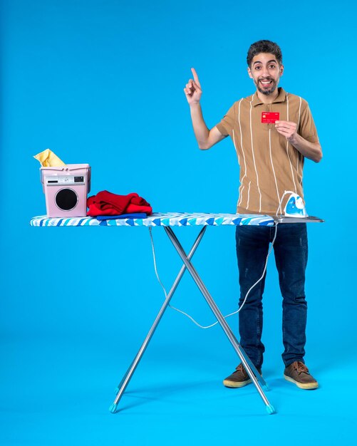 Photo front view young male behind ironing board holding bank card on blue background laundry housework washing machine iron money color clean