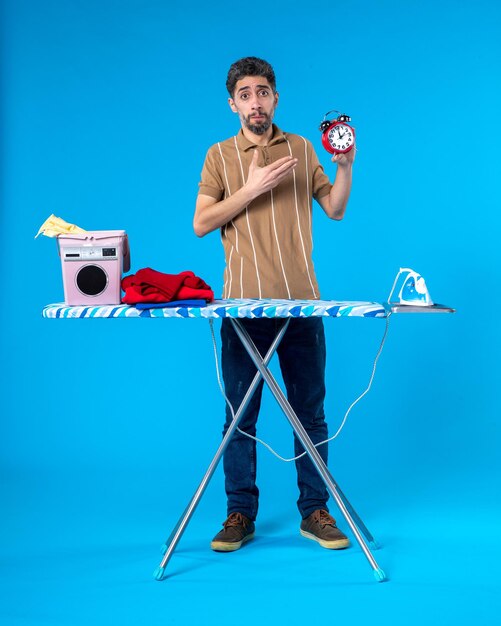 front view young male behind ironing board holding clocks on a blue background time housework washing machine clean laundry color iron