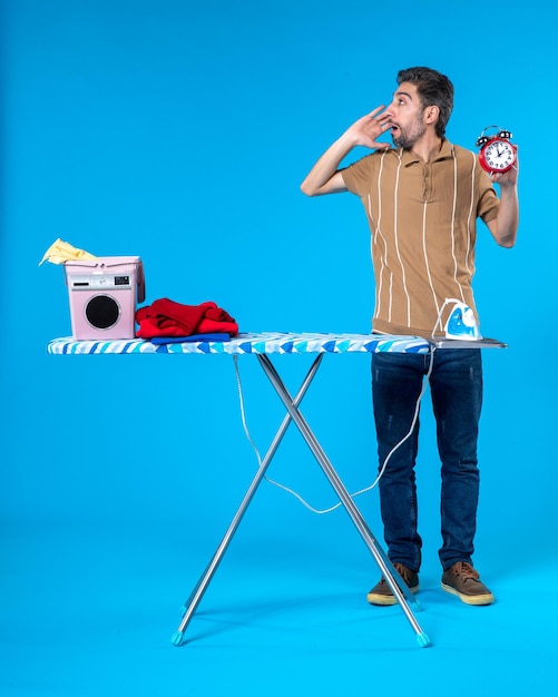 front view young male behind ironing board holding clocks on blue background washing machine clean laundry iron time color