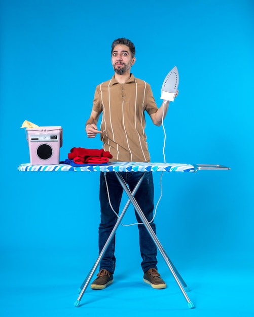 Photo front view young male behind ironing board holding iron on a blue background color housework clean laundry wash machine man emotion