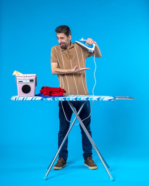 front view young male behind ironing board holding iron on blue background color housework clean machine emotion man laundry