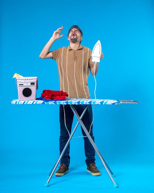 front view young male behind ironing board holding iron on blue background color housework clean machine man laundry emotion