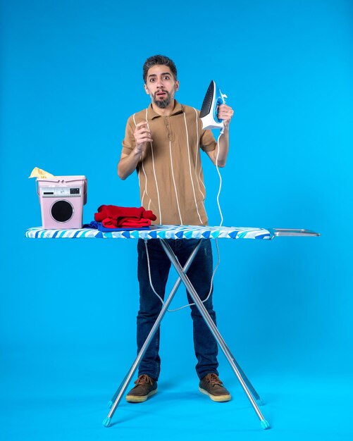 Photo front view young male behind ironing board holding iron on blue background color housework clean machine wash man laundry emotion