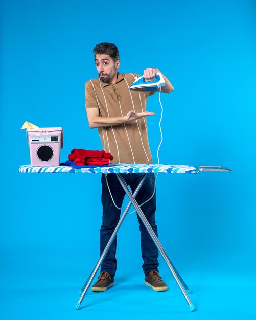 Front view young male behind ironing board holding iron on blue\
background color housework clean wash emotion man laundry