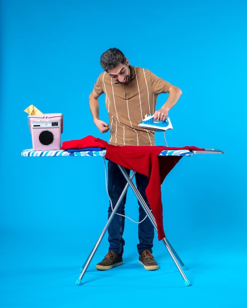 Front view young male behind ironing board preparing to iron on a blue background clean laundry washing machine color housework