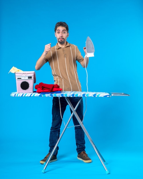 Front view young male behind ironing board taking iron on the\
blue background color housework laundry man clean washing machine\
emotion