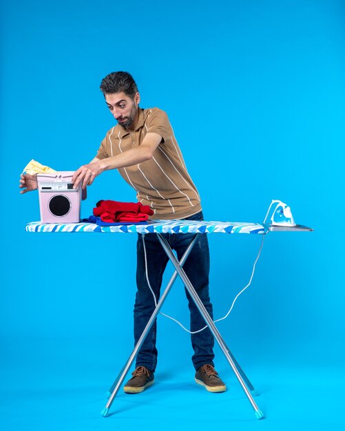 Front view young male behind ironing board taking off clean towels on a blue background color housework laundry man emotion clean wash machine